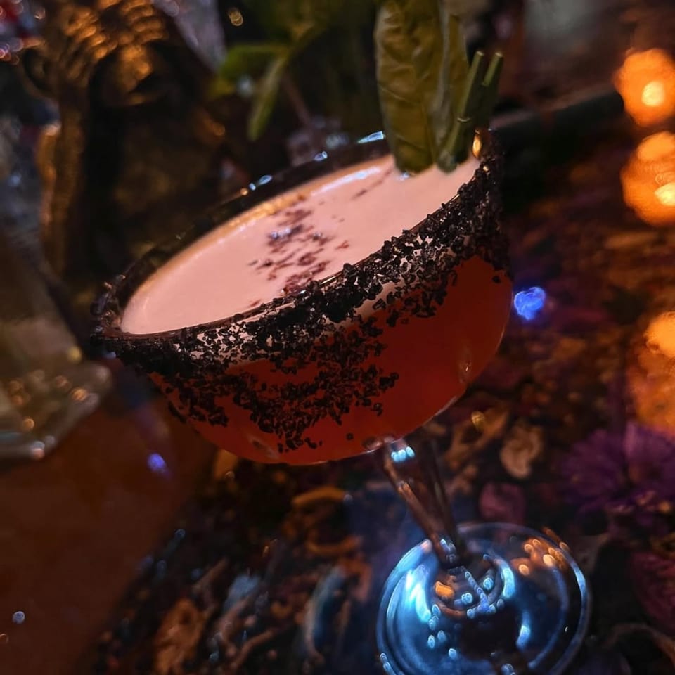 A red cocktail with a black sugar rim and green garnish sits on a dark, patterned surface.