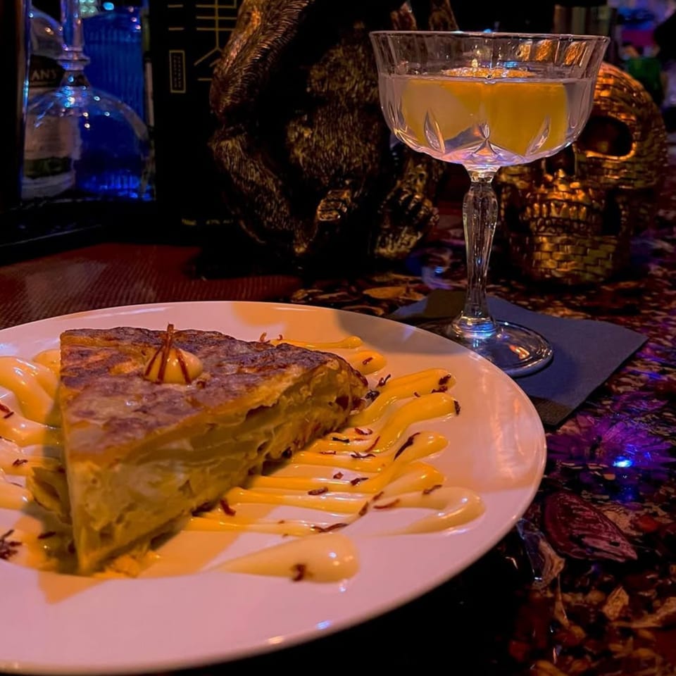 A slice of Spanish tortilla with sauce on a white plate, next to a cocktail and a golden skull.